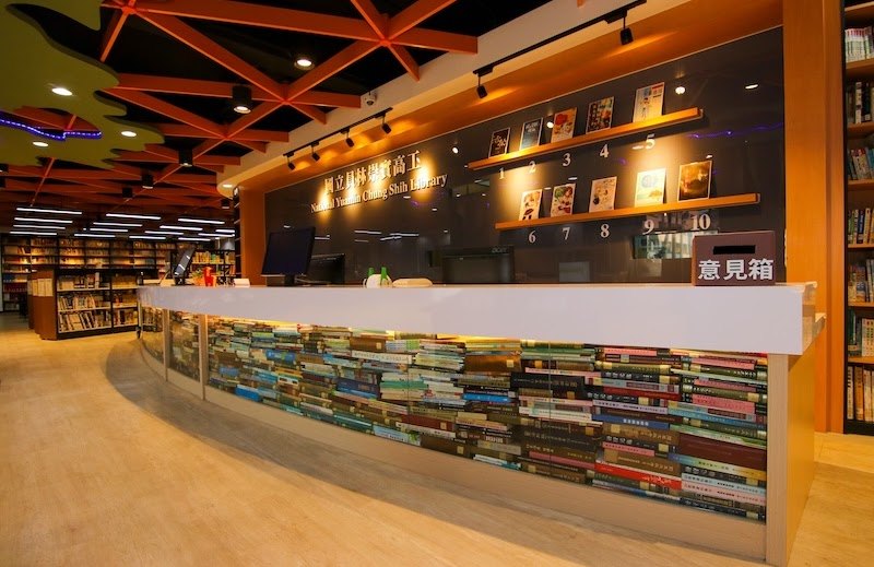 低矮的白色流通櫃台，台面下是整排透明展示櫥窗，後方牆面是館長推薦書展示架 The low white circulation desk with a run of glass display windows beneath it and the librarian's picks displayed on the wall behind