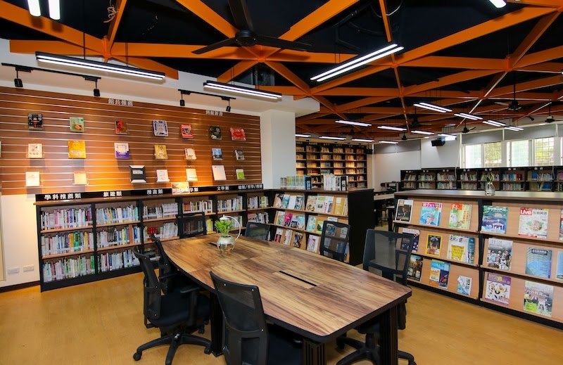 木紋波浪造型長桌配上工業風座椅，兩側是雜誌與新書展示架 A wood-grain wavy table with industrial-style chairs, magazine and new-book racks on both sides