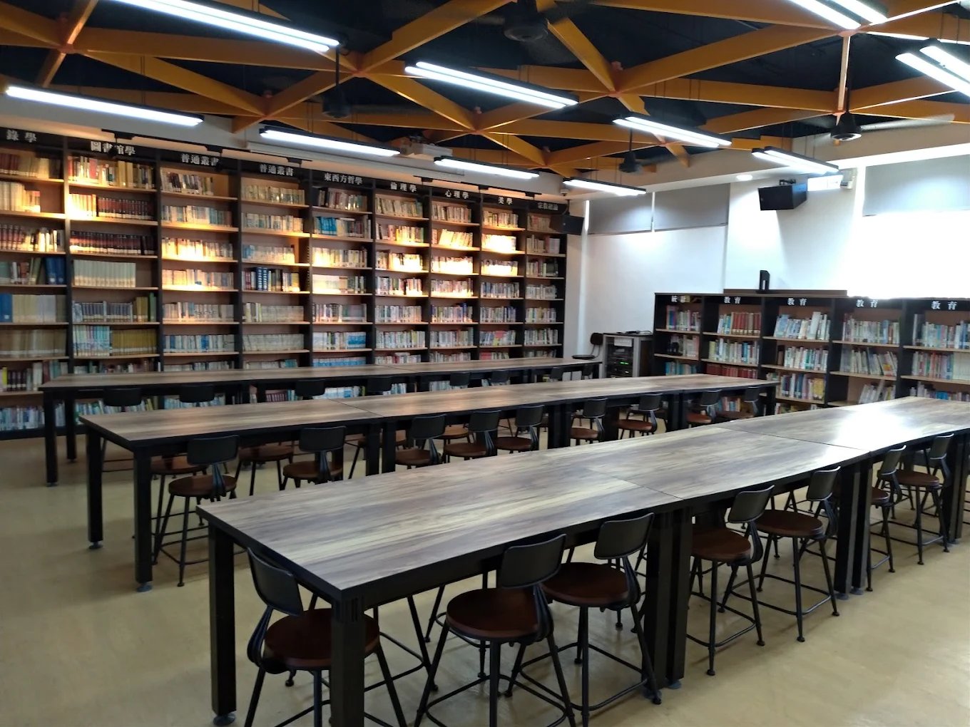 崇實圖書館的閱覽區，兩排長桌與高腳椅，後方是一整面依類別分區的書架 The reading room at the ChungShih library, two long shared tables with stools, subject-sorted shelves filling the wall behind