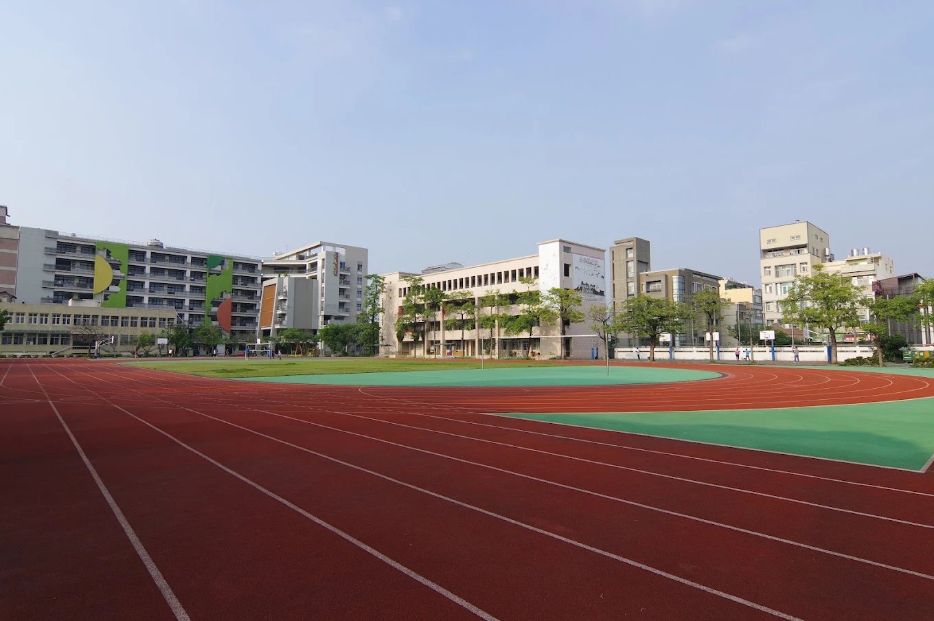 崇實高工的操場，紅色 PU 跑道環繞綠色球場，後方是一整排校舍 The sports field at ChungShih, a red track around a green infield with the line of school buildings behind